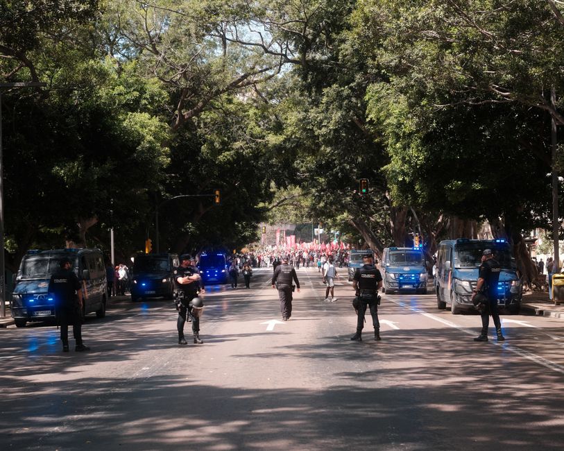 Agentes de policía bloquean una carretera durante una protesta en Málaga, España, bajo la sombra de los árboles en un día soleado.