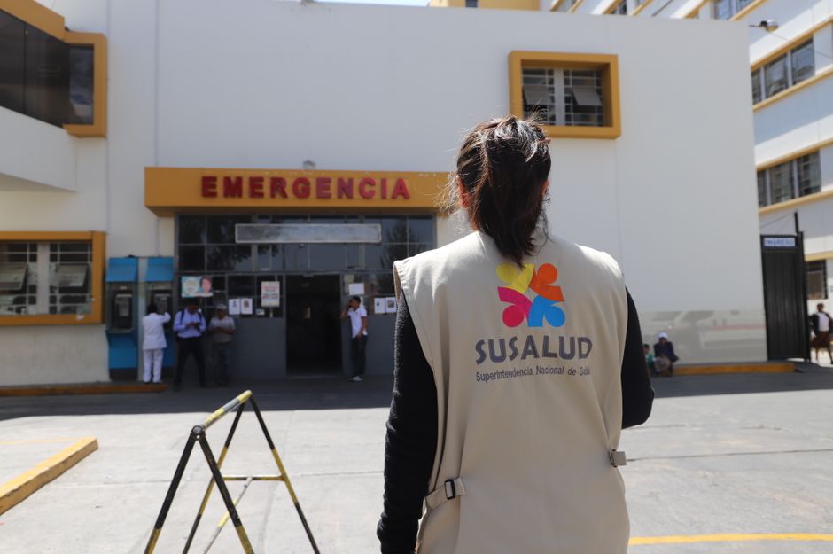 Un oficial de SUSALUD afuera de la entrada de emergencia de un hospital durante el día.