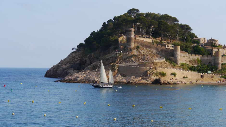 Vista escénica del castillo histórico de Tossa de Mar con vista al Mediterráneo y un velero en primer plano.
