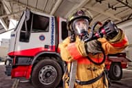 Bombero con equipo de protección sosteniendo una manguera cerca de un camión de bomberos en un entorno de garaje.