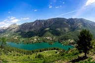 Vista aérea de un pintoresco lago de montaña rodeado de exuberante vegetación y majestuosos picos bajo un claro cielo azul en Valencia, España.