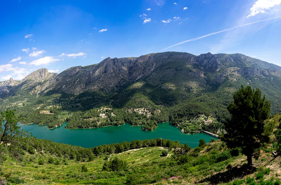 Vista aérea de un pintoresco lago de montaña rodeado de exuberante vegetación y majestuosos picos bajo un claro cielo azul en Valencia, España.