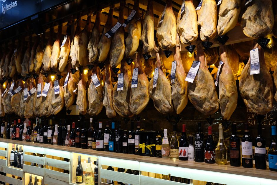 Una exhibición vibrante de jamón español colgado con botellas de vino en un mercado de Barcelona.